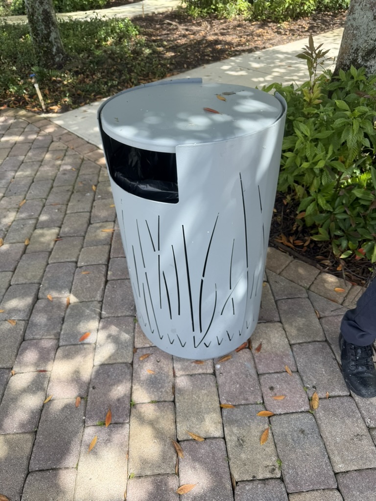 A current outdoor waste bin at TAO Sawgrass — a faded grey cylindrical can with grass-blade cut-out pattern, exposed black plastic liner visible through the opening, sitting on the paver walkway.