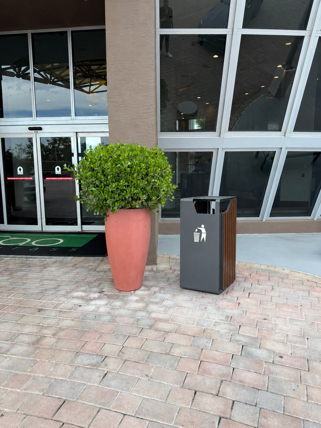 Rendering of the new dark anthracite wood-slat waste bin in place at the main lobby entrance — to the right of the large terracotta planter with topiary, beside the automatic sliding glass doors and the green TAO Sawgrass entry mat.