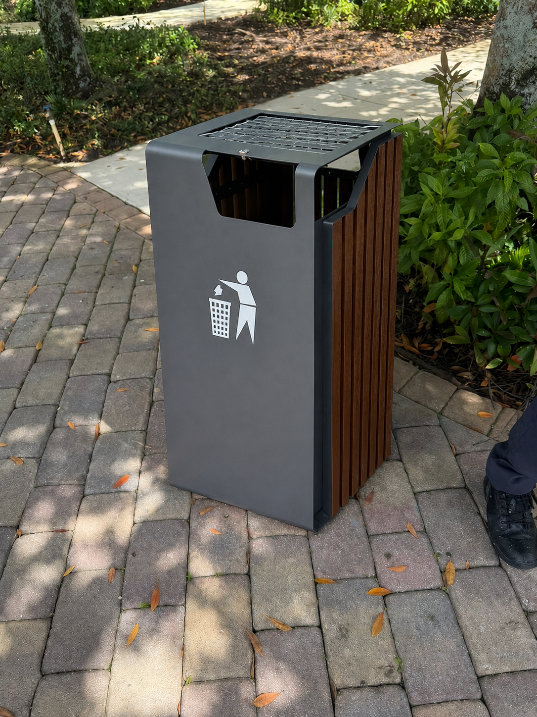 Detail view of the proposed waste bin — dark anthracite powder-coated steel body, vertical warm-tone wood slat side panels, top mesh grate for ash and small litter, side opening with hidden plastic liner, and a tidyman pictogram on the front face.