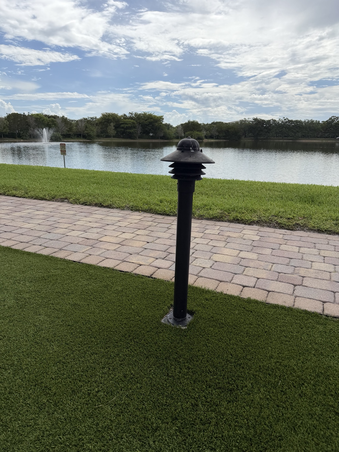 The lakeside walkway today — a weathered three-tier pagoda-style black bollard, paint chipped and faded, standing on a black metal base in the turf next to the paver path with the lake and fountain behind.