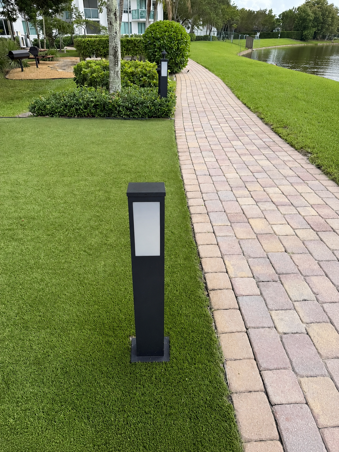 View along a paver walkway curving toward the building with the lake on the right, two of the new dark frosted-lens bollards spaced along the path edge in the manicured turf, creating a uniform path-lighting rhythm.