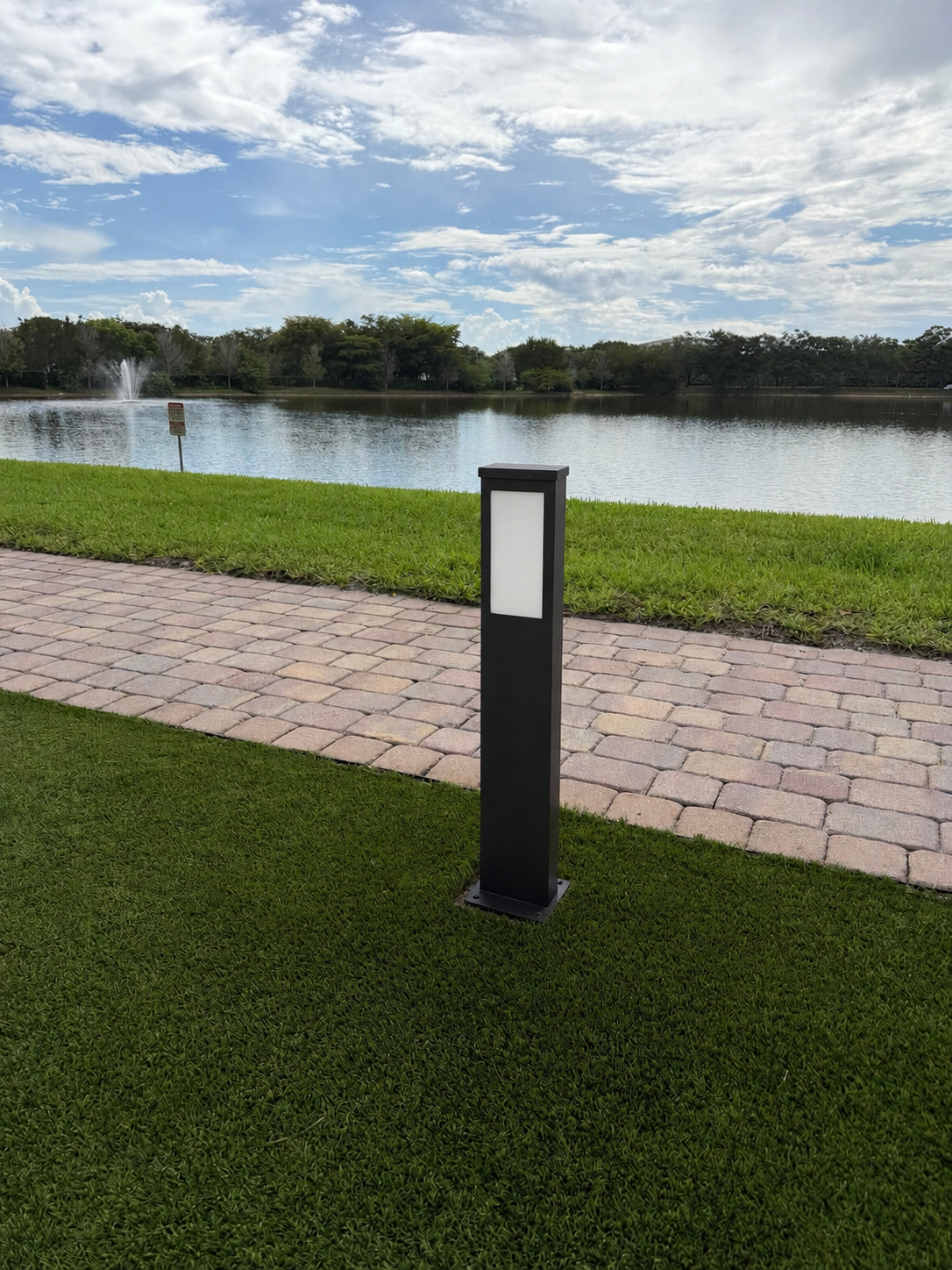 Rendering of the new modern bollard at the same lakeside spot — a rectangular dark anthracite aluminum post with a tall frosted-glass lens panel emitting a warm, even glow, on a low metal mounting plate in the turf next to the paver path.